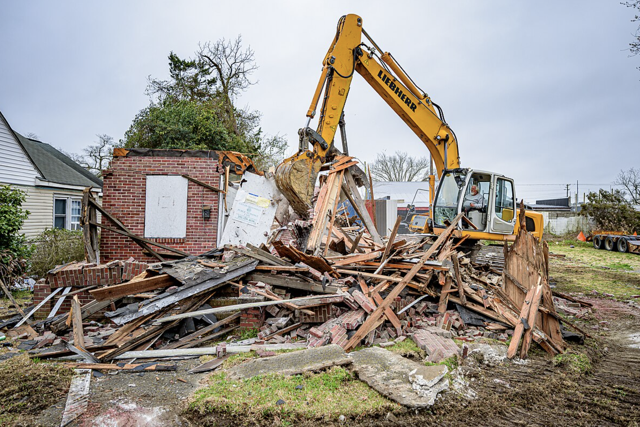 A yellow excavator is actively digging near a house under construction.