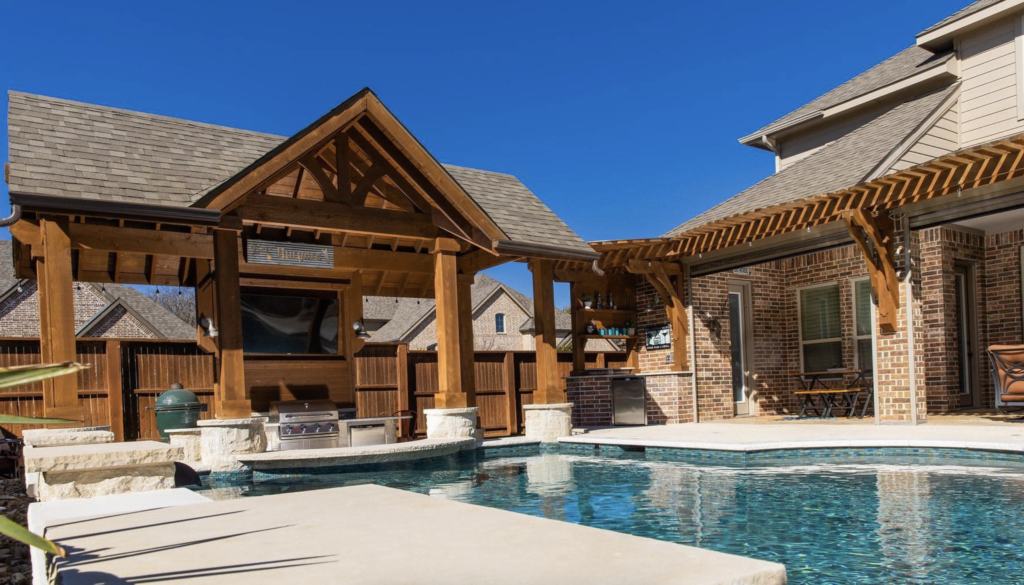 Outdoor scene with a serene pool surrounded by a brick house and wooden pergola. The clear blue sky adds to the bright, inviting atmosphere.