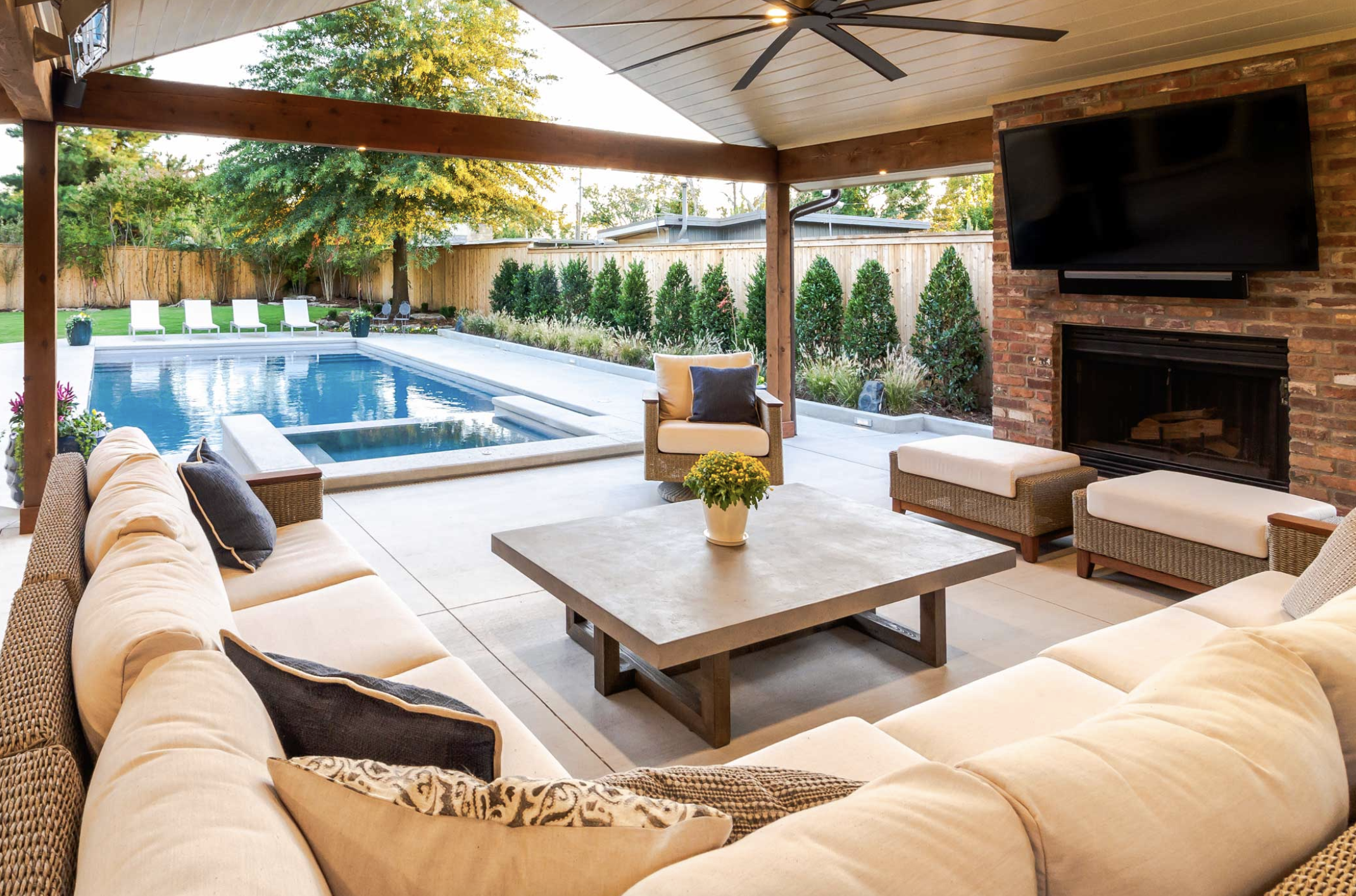 Outdoor patio with beige couches around a large table, facing a pool. There's a fireplace and TV on a brick wall. The setting is cozy and inviting.