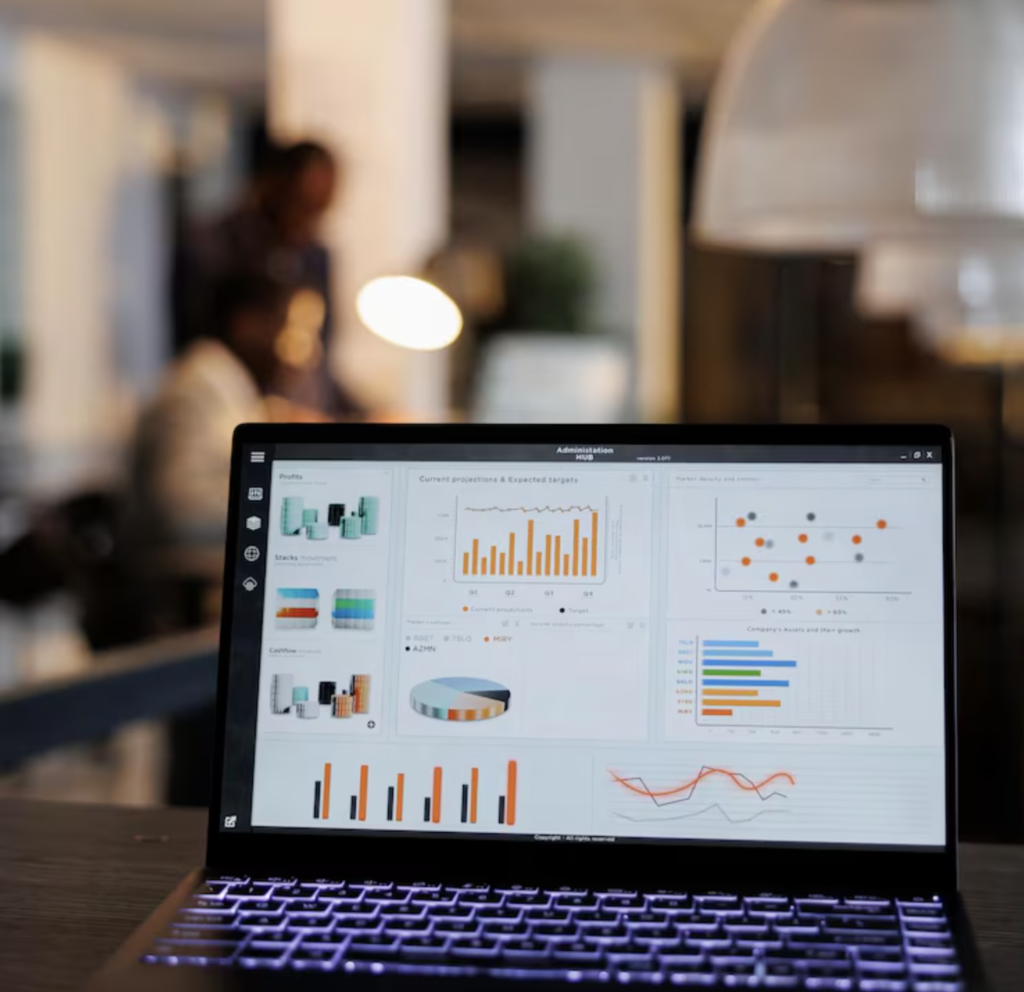 A laptop screen displaying colorful data visualizations, such as bar and pie charts, in a dimly lit office. Blurry figures are seen in the background.